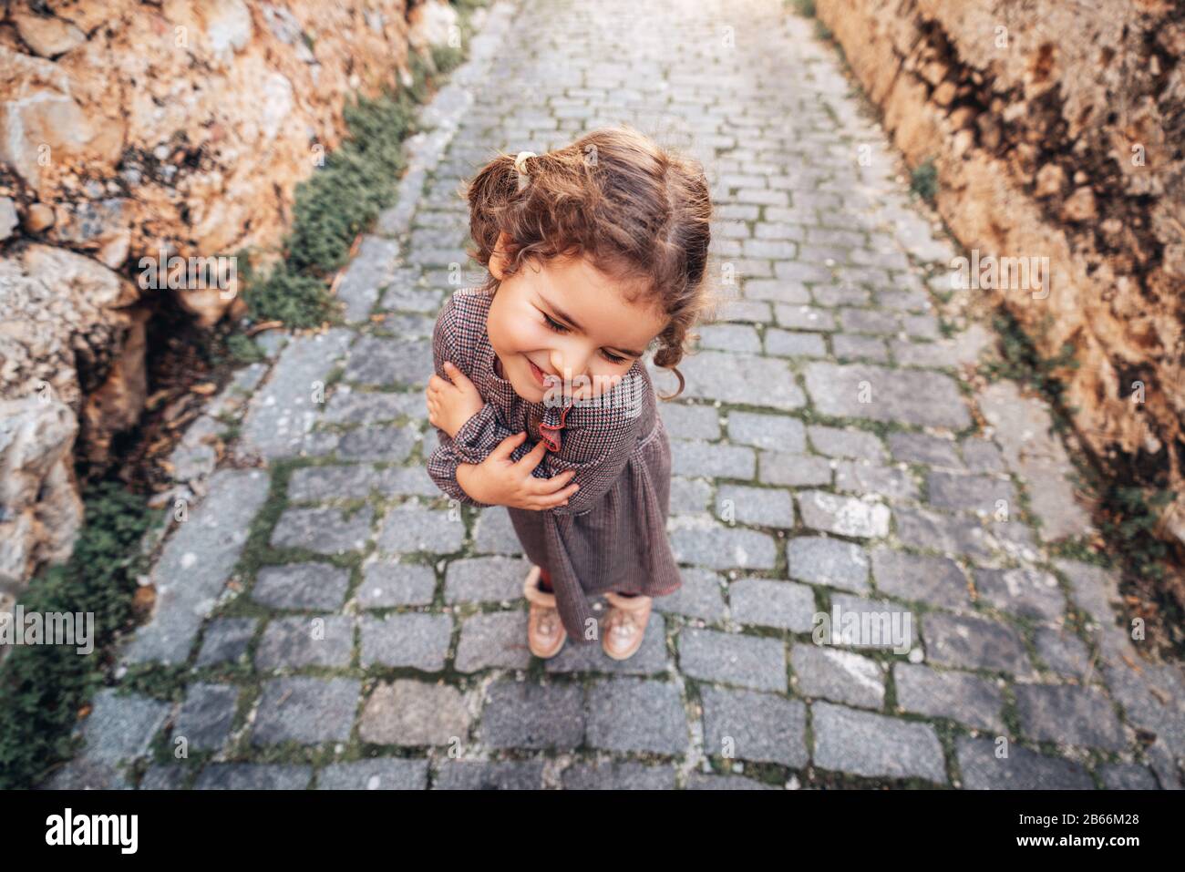 Pretty baby girl hold arms closed with a shy gesture and walk on a footpath on a medieval alley Stock Photo
