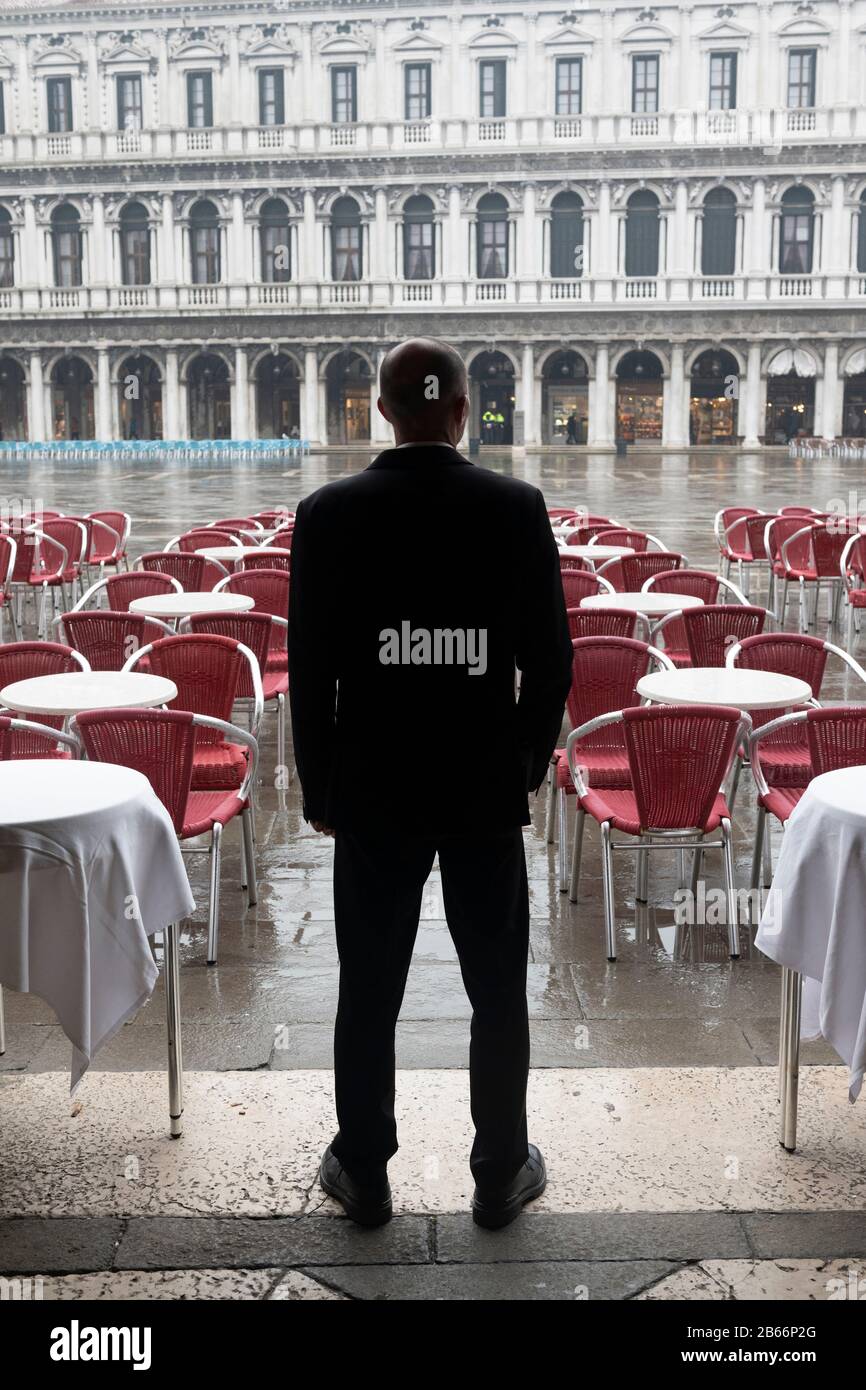 Venice, Piazza San Marco  March 2nd.. 2020: A restaurant owner stands outside his premises looking across an empty St. Marks Square, one of the most popular locations in Venice for tourists. Venice streets deserted due to Coronavirus pandemic. Piazza's and streets are empty apart from a handful of tourists and locals going about their daily business. Many tourists cancel their visit for fear of catching the virus. Tourists from Japan and other far eastern countries like Taiwan  often wear surgical masks as a preventative measure. Piazza San Marco (St. Marks Square, one of the most popular tour Stock Photo