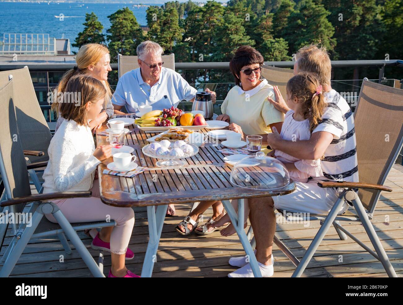 Big happy family having breakfast outdoors on terrace together, sitting around table, drinking coffee. Beautiful sea view, warm summer morning. Family Stock Photo