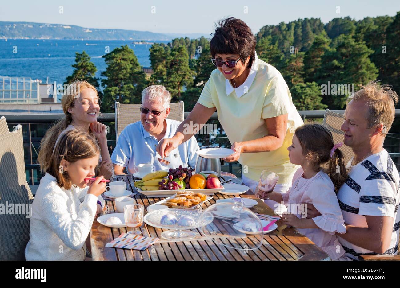 Big happy family having breakfast outdoors on terrace together, sitting around table, drinking coffee. Beautiful sea view, warm summer morning. Family Stock Photo