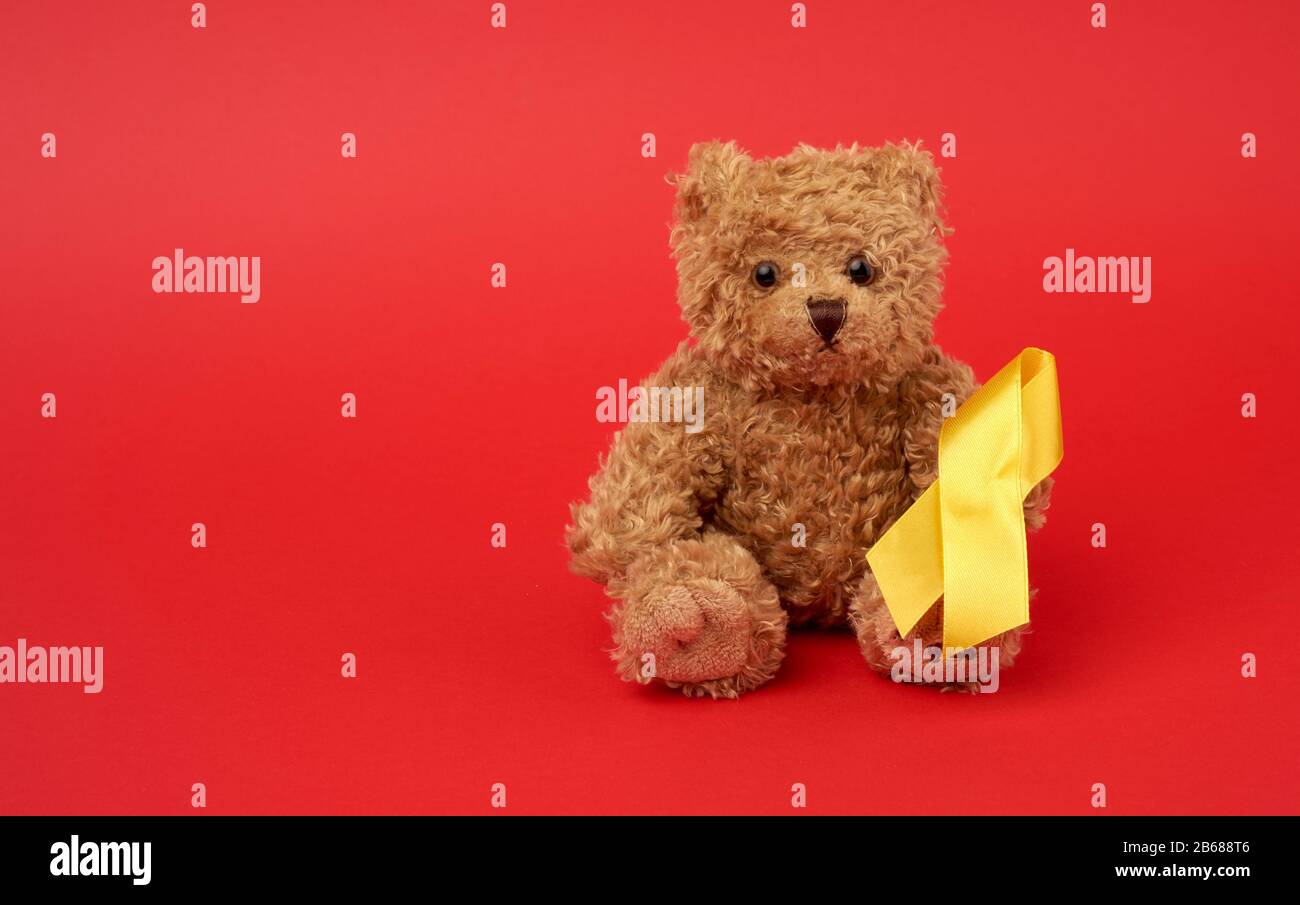 Cute brown teddy bear holds a silk yellow ribbon in the shape of a loop on a red background, concept of the fight against childhood cancer, problem of Stock Photo