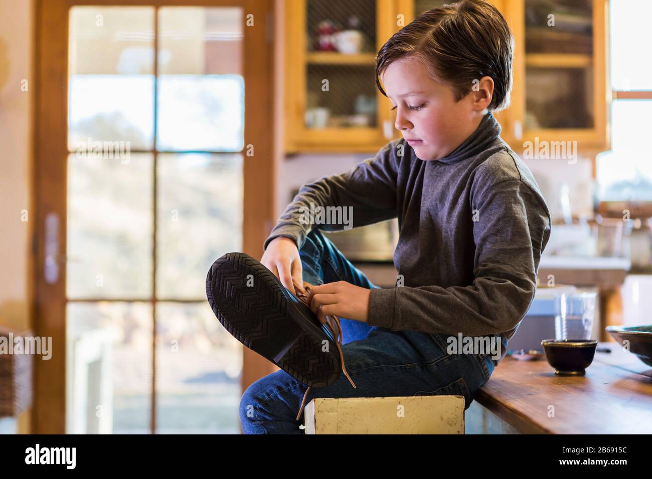 A six year old boy putting his boots on, sitting on a high stool. Stock Photo