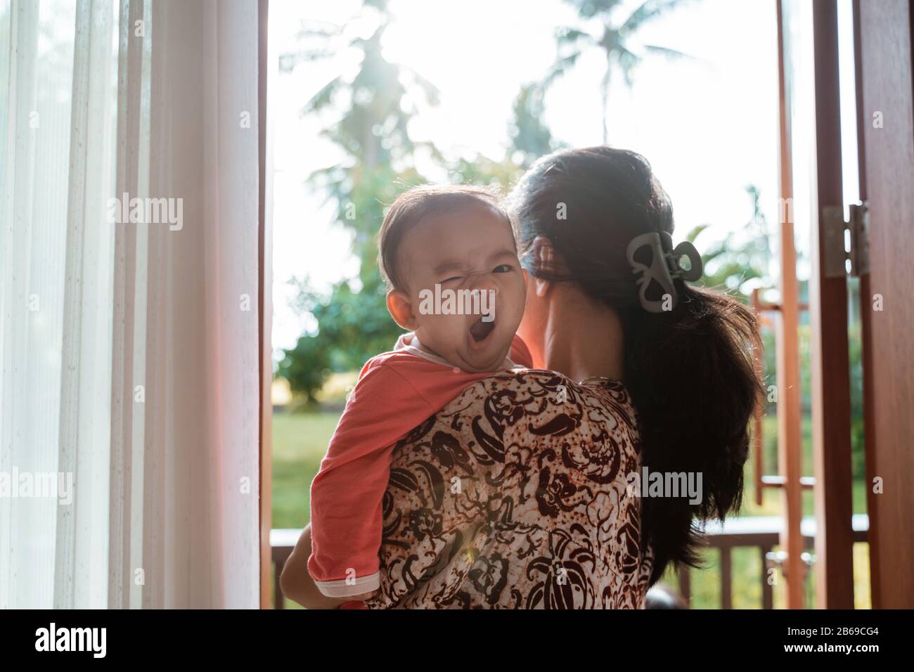 asian mother holding a sleepy baby girl while standing in front of the door Stock Photo