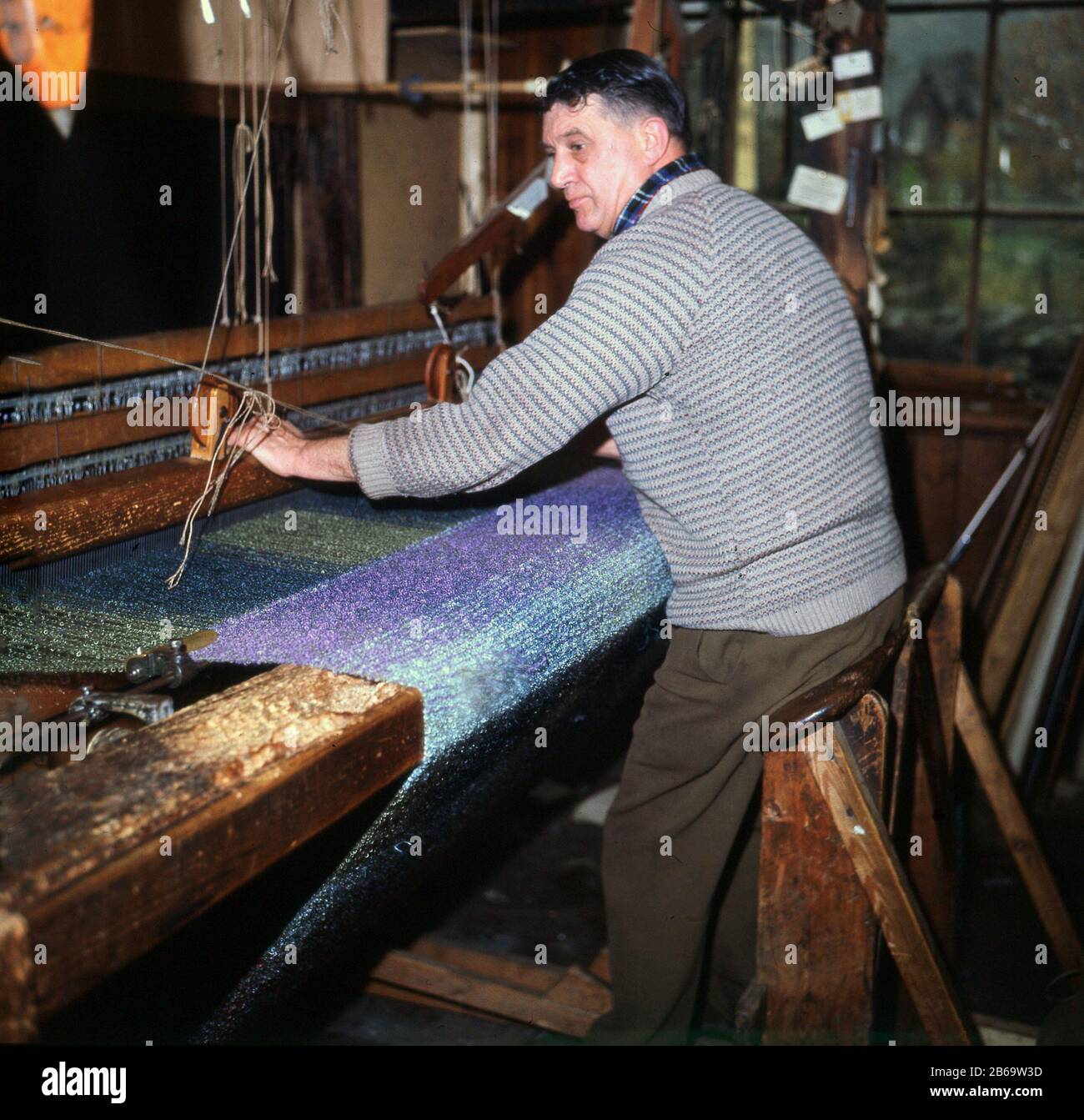 1960s, a man weaving mohair cloth by hand on a wooden loom, Grasmere, Cumbria. A silk-life fabric or yarn, mohair is made from the long hair of the Angora goat and as can be seen takes dyes well, to produce vibrant colourful garments. Stock Photo