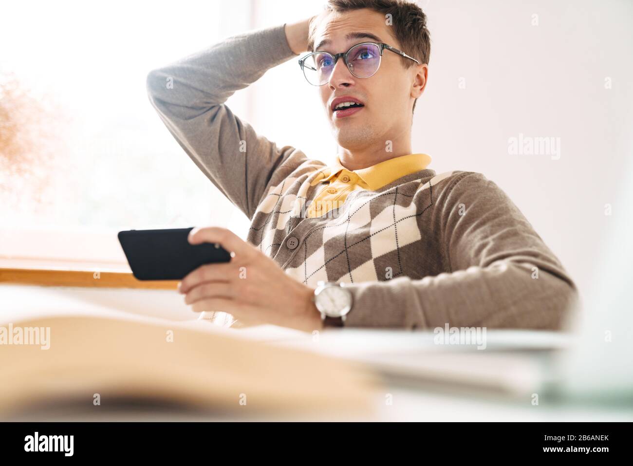 Picture of a young shocked guy student sit at the table indoors using mobile phone. Stock Photo