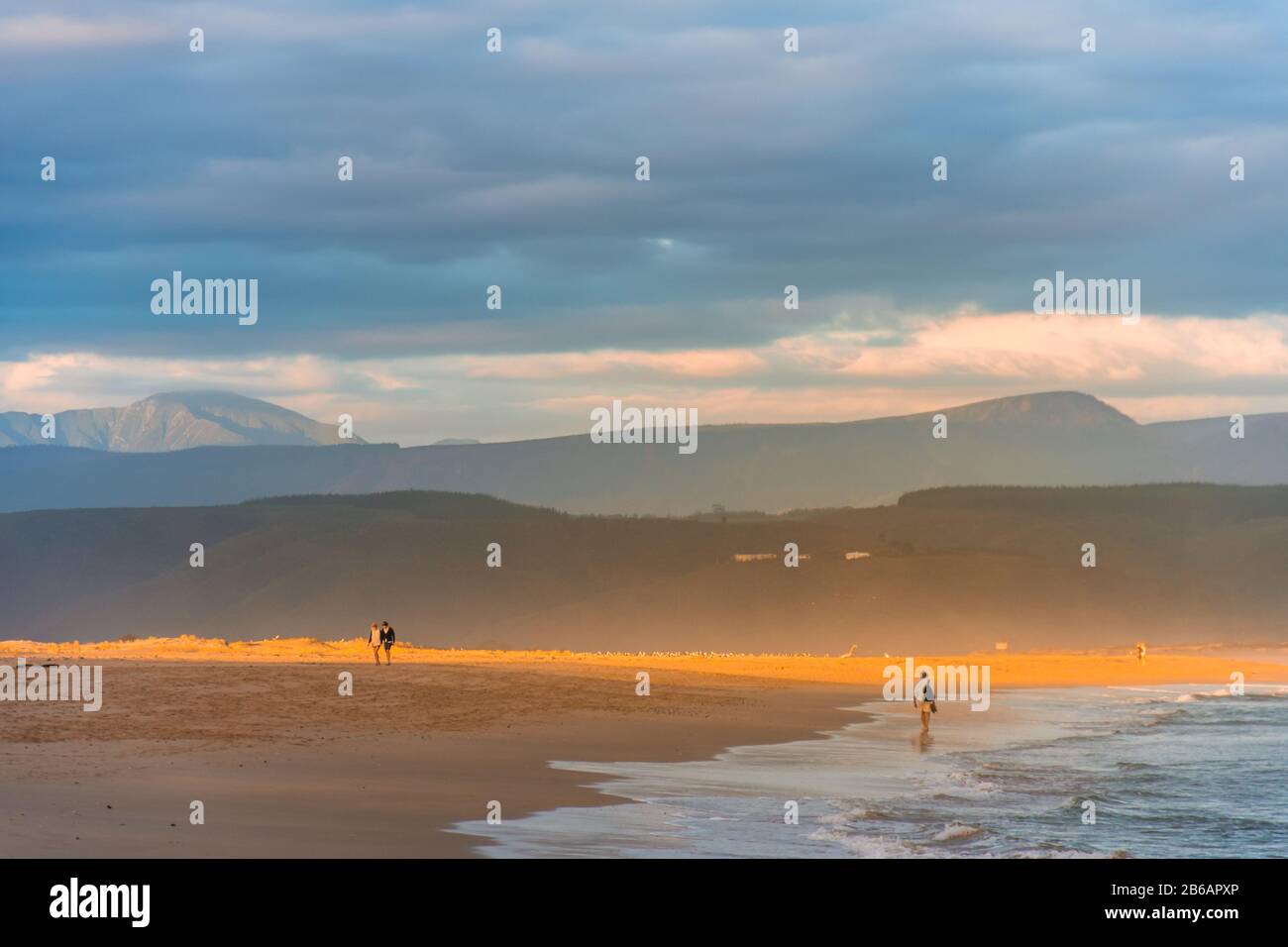 A streak of golden evening light falling on the Plettenberg Bay beach at sunset, with mountains in the distance and people walking on the beach. Garde Stock Photo