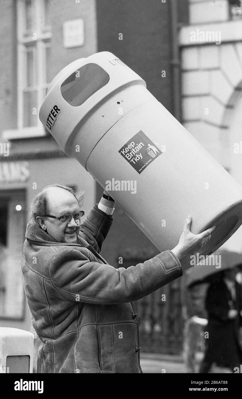 1987, historical, in a high street, a man in a sheepskin coat lifting a plastic litter bin, marking the start of the 'Keep Britain Tidy' campaign, York, England, UK. Stock Photo