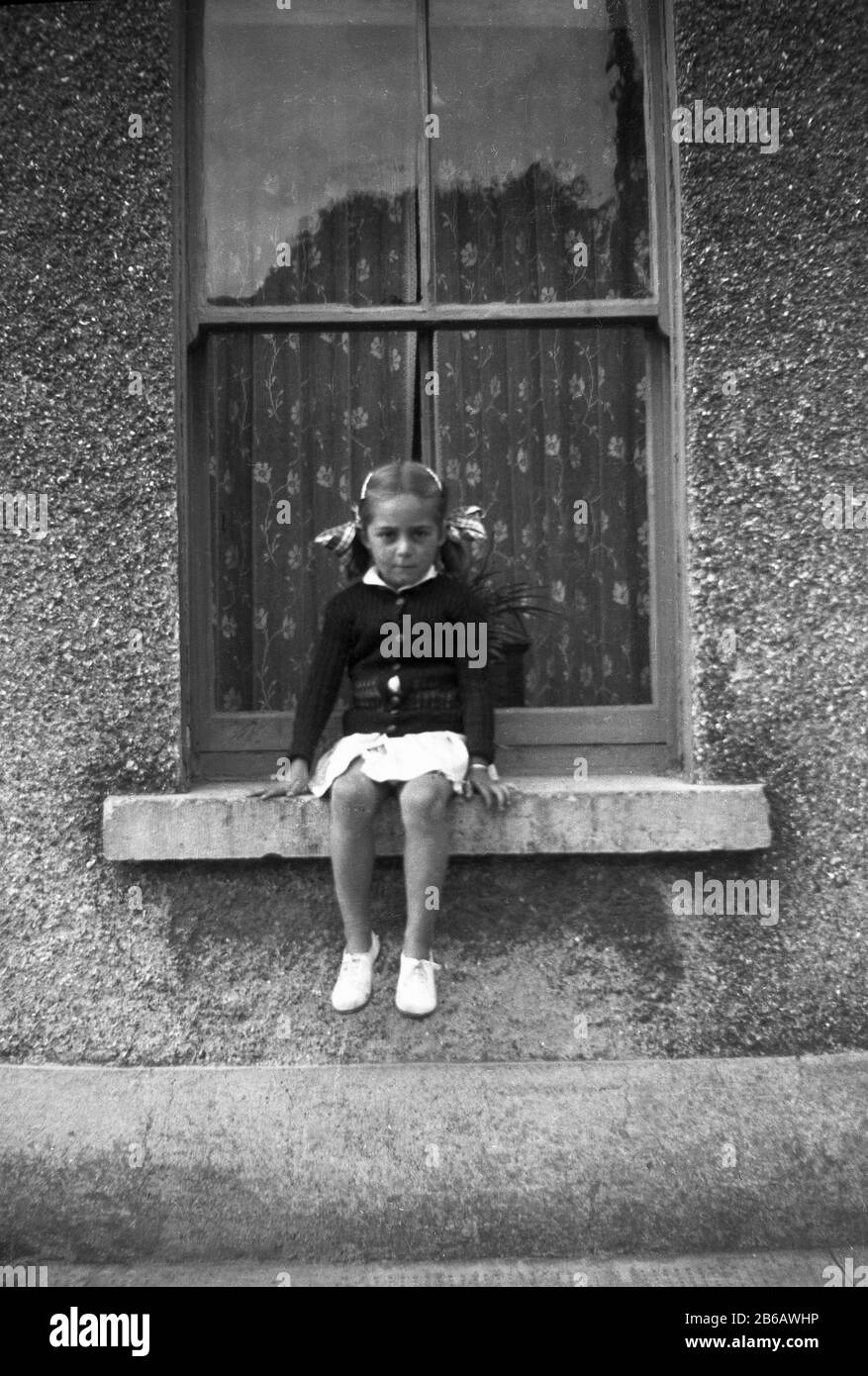 1950s, historical, young girl sitting outside on a window ledge of an old wooden sash window of a pebble dash house, England, UK. Stock Photo