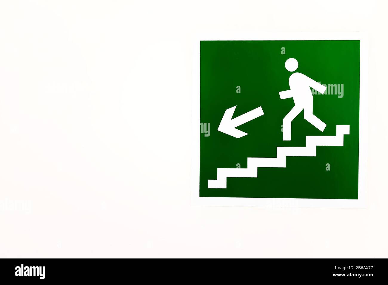 A green sign down the stairs on an empty bright wall. DIRECTION TO THE EMERGENCY EXIT ON THE LADDER DOWN Stock Photo