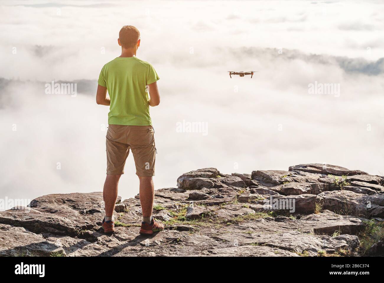 Young Man navigating a flying drone at fog and clouds, standing on top of  the mountains at sunset Stock Photo - Alamy