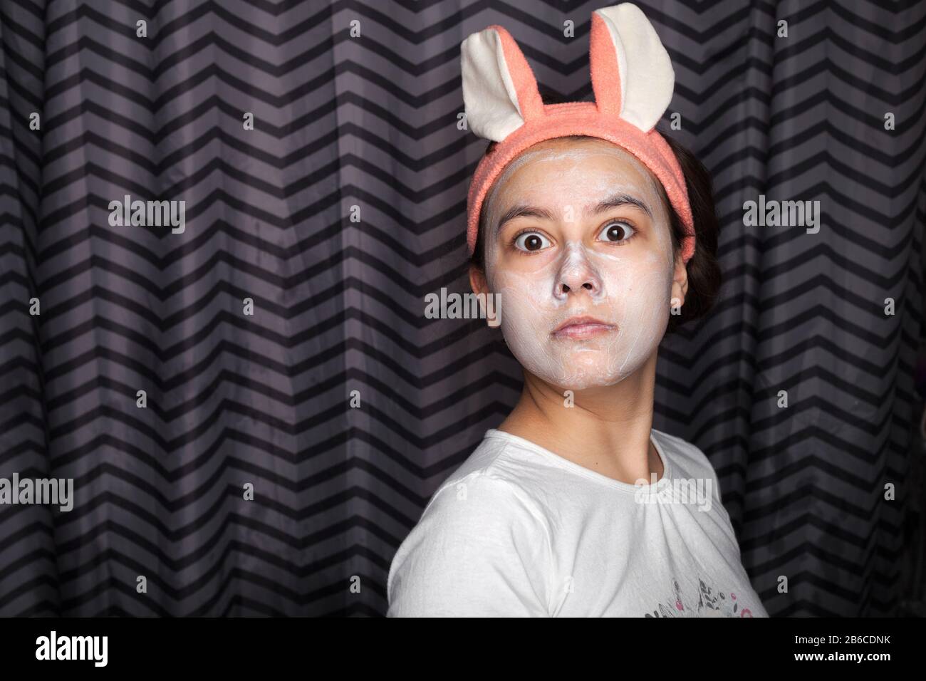 A happy girl with cosmetic clay or mud on her face, standing in the bathroom, with rabbit ears on her head and grimaces funnyly. The concept of health and beauty. Stock Photo