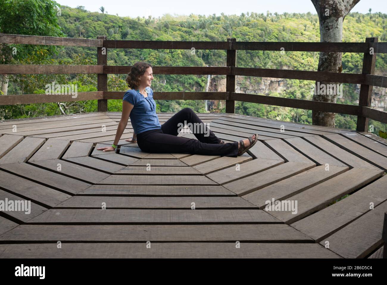 Woman sitting relaxed on a wooden deck with octagonal shape, looking at the natural view of Ubajara National park with forest and waterfall on the bac Stock Photo