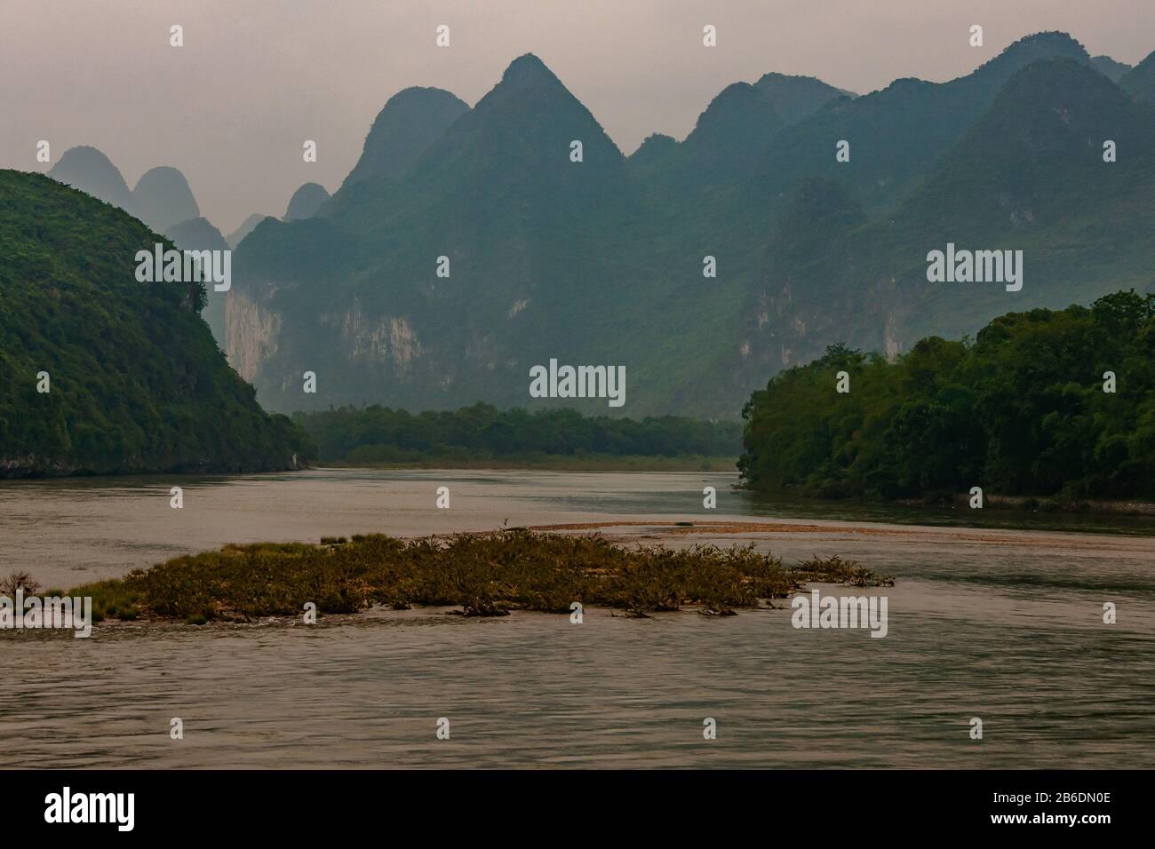 Guilin, China - May 10, 2010: Along Li River. Landscape with green forested karst mountain and cliffs under foggy sky. Brown water and marsh land up f Stock Photo