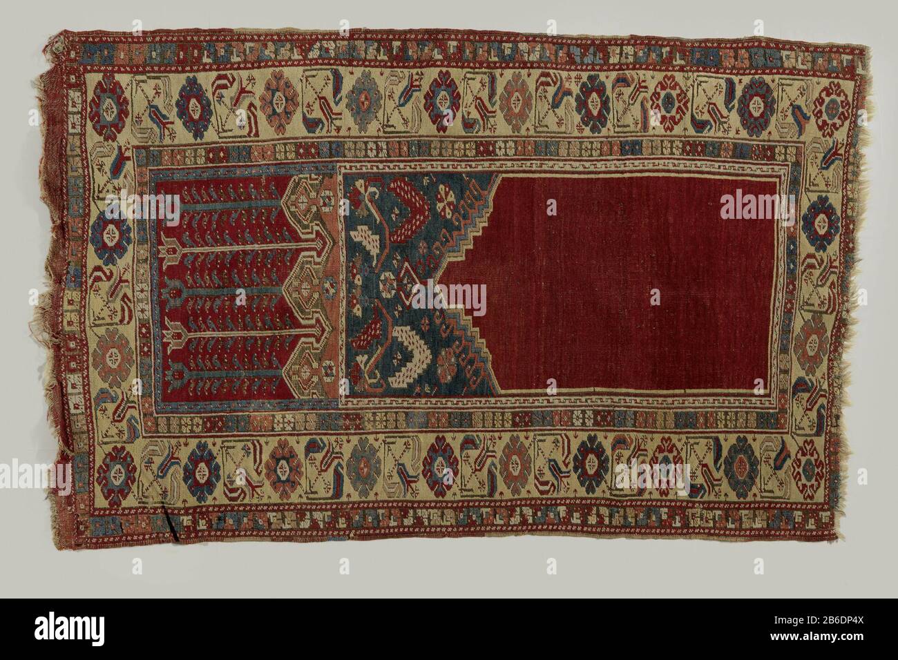 Prayer rug with upper transverse panel, Ladik Bidkleed met bovendwarspaneel, LADIK. Middenveld: effen rode mihrab met een in het midden verhoogde trapgeveltop. Blauwe zwikken met veelkleurige sterbloemen en getande lancetbladeren aan een liggende S-rank. Een kantelenrand is in roze met witte omegalijn, ingrijpend in een rood bovenpaneel, Stock Photo