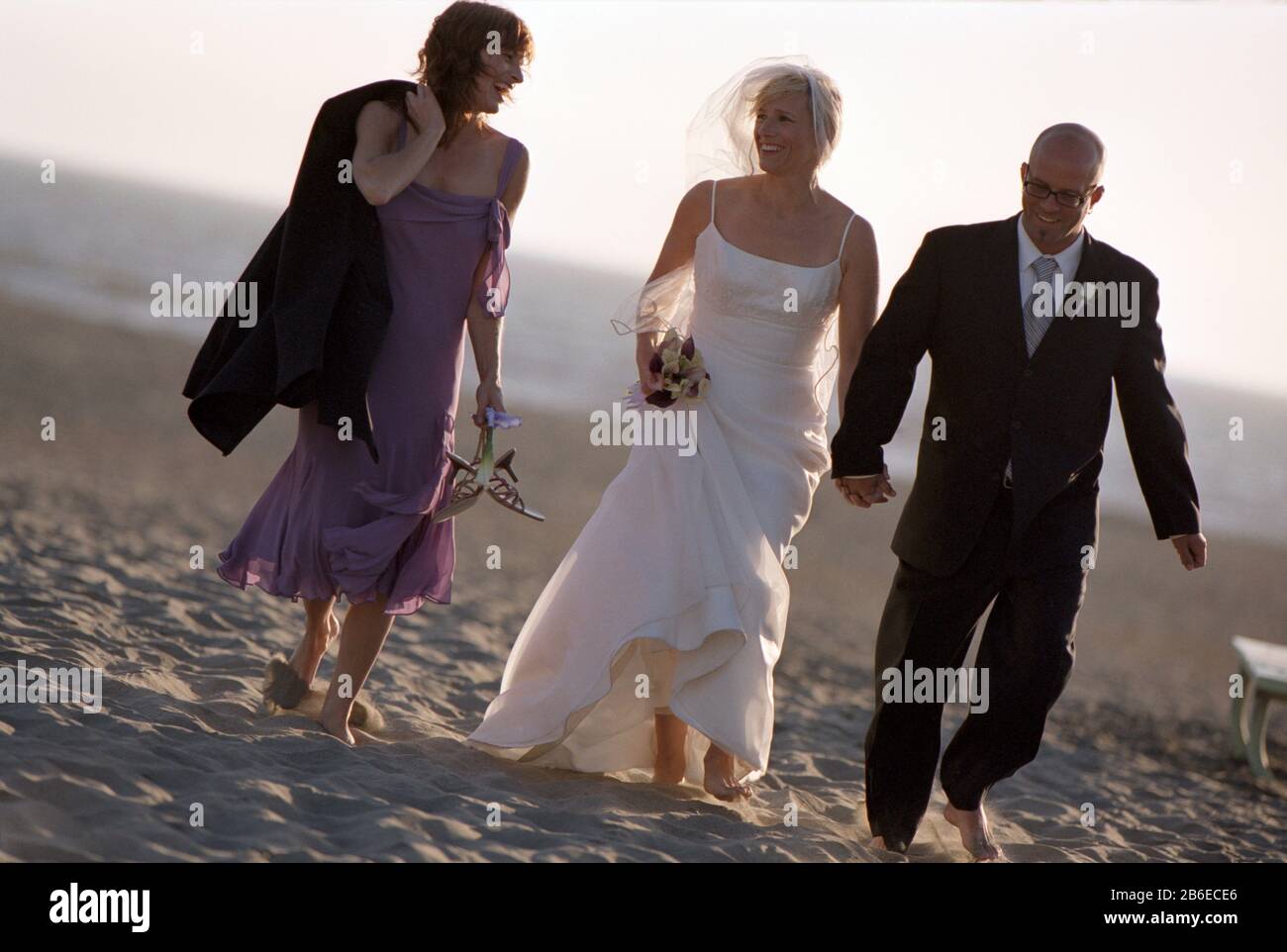 Smiling bride and groom walking along a sandy beach with one of their bridesmaids. Stock Photo