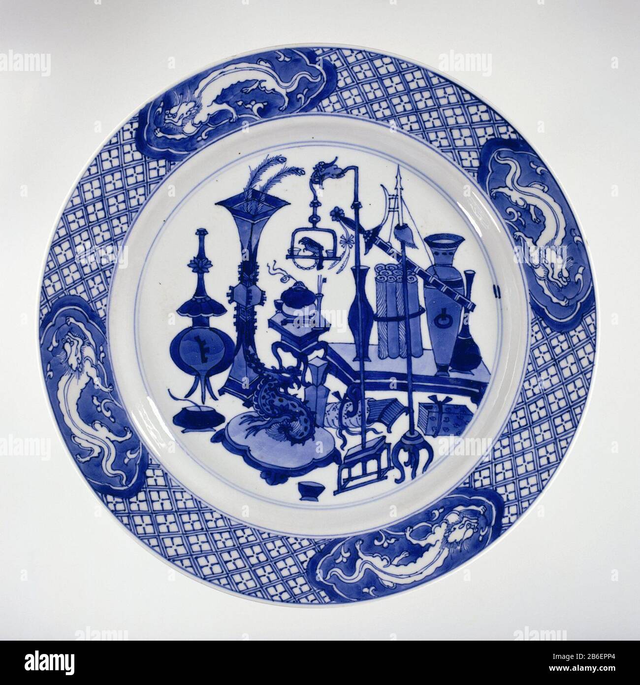 Plate with antiques and dragons Bord van porselein met opstaande wand en schuine rand, beschilderd in onderglazuur blauw. Op het plat antiquiteiten (vazen, pauwenveren, boeken, rollen, Wie: rrookbrander) en wonderlijke objecten