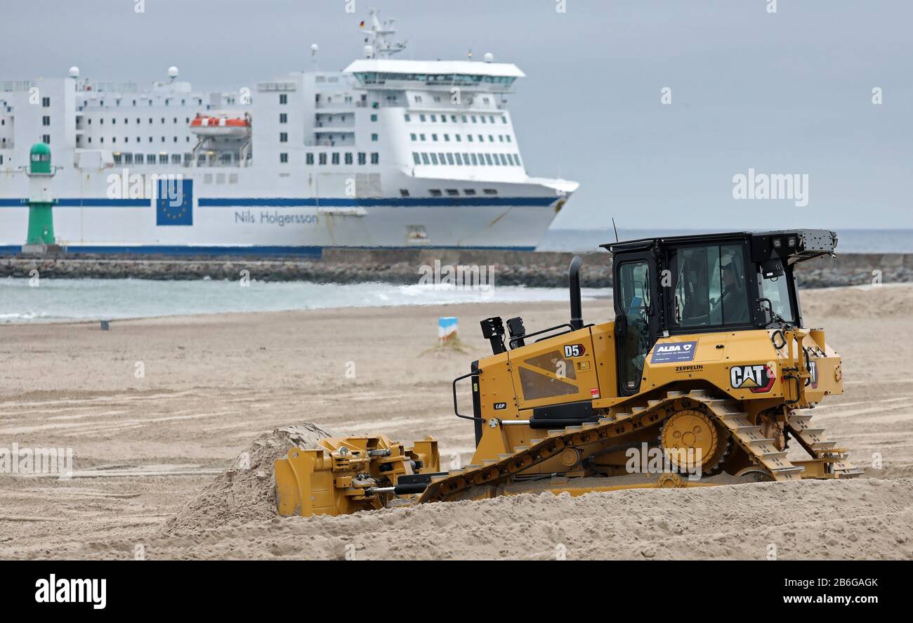 10 March 2020, Mecklenburg-Western Pomerania, Warnemünde: A bulldozer levels the Baltic Sea beach and fills up the areas with sand that have been affected by the autumn and winter storms. In the background, the ferry 'Nils Holgersson' of the shipping company TT-Line from Trelleborg (Sweden) enters the port. On the beach the first preparations for the new season have started. Photo: Bernd Wüstneck/dpa-Zentralbild/dpa Stock Photo