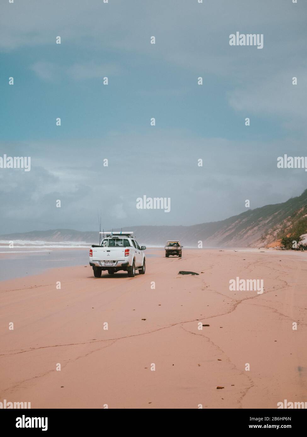 Two cars driving on the shore of the beach with mountains in the background in Australia Stock Photo
