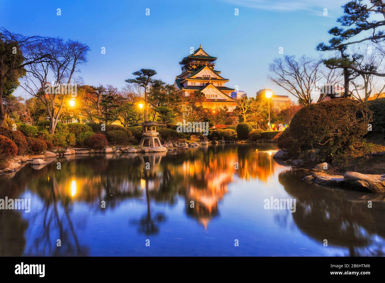 Still pond in traditional japanese garden of Osaka city with historic tower and street lights reflecting at sunrise. Stock Photo