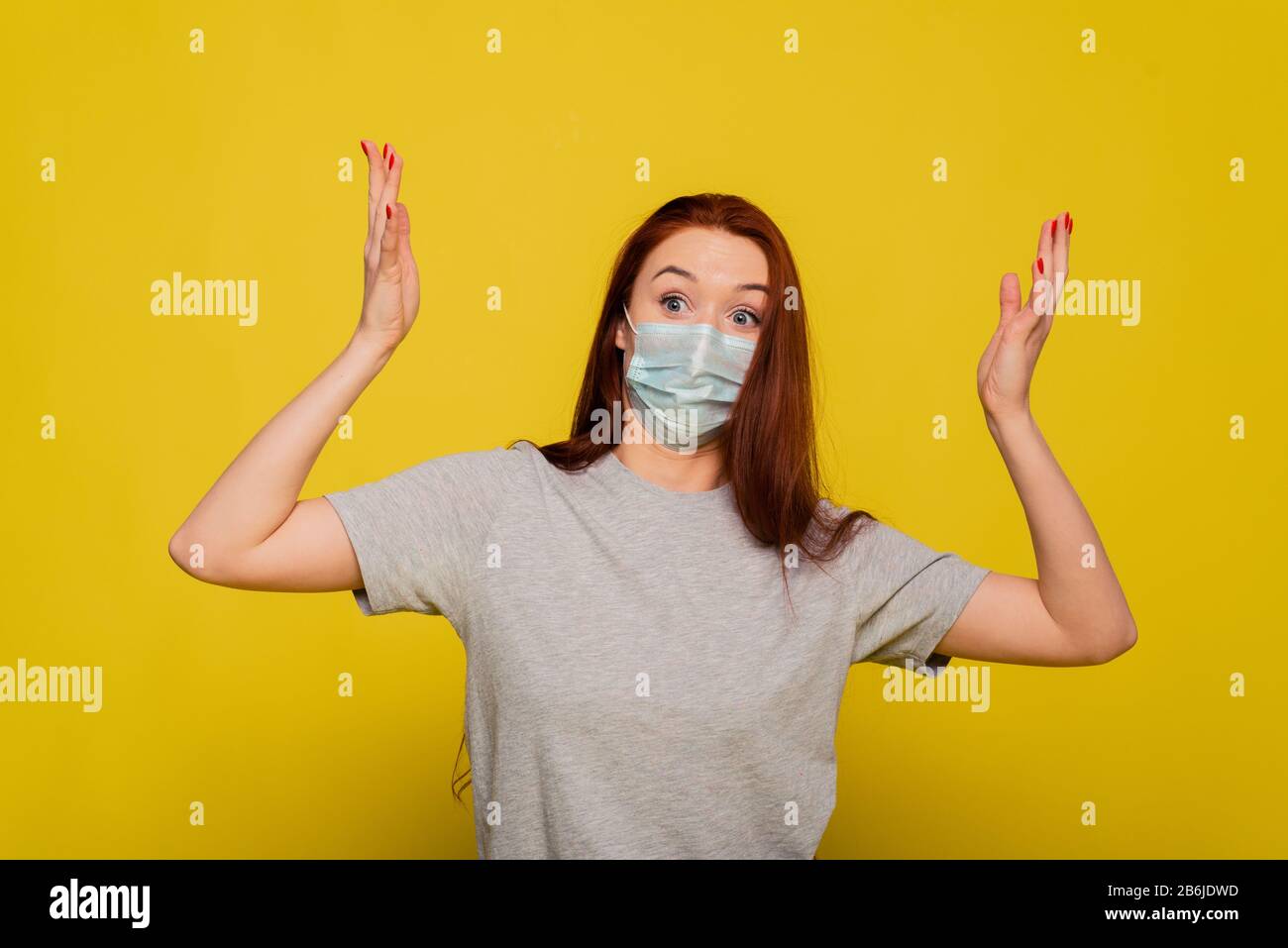 A woman clings to her head and experiences fear, shock from the spread of the disease. Red-haired beautiful girl in a protective mask on a yellow background screams from a coronavirus. The concept of protection and prevention. Stock Photo
