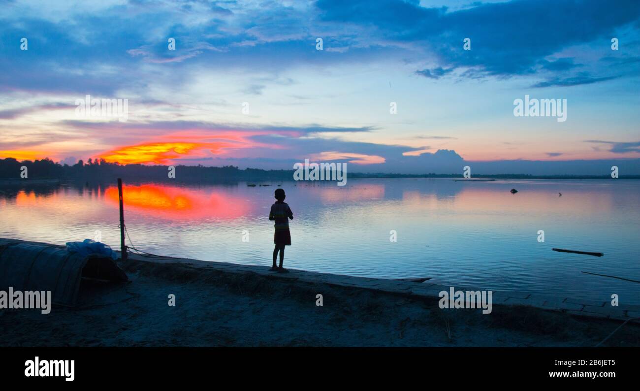 A boy is standing alone on the bank of a river in the evening light at Northern Bangladesh. The beauty of the moment felling the boy like an anger. Stock Photo
