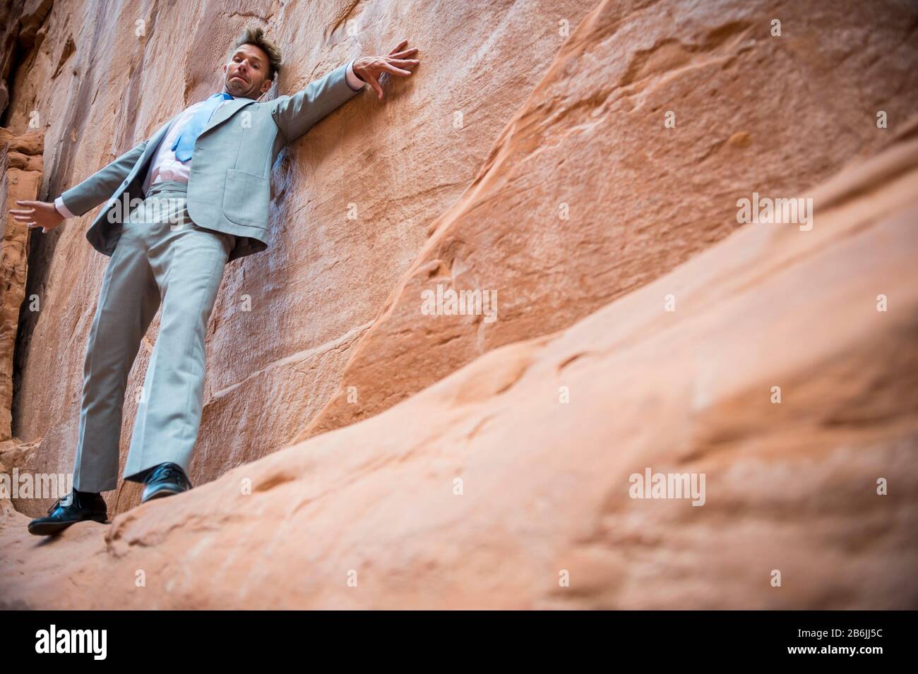 Nervous businessman clinging to a cliff face while balancing on a narrow ledge in a red rock canyon Stock Photo