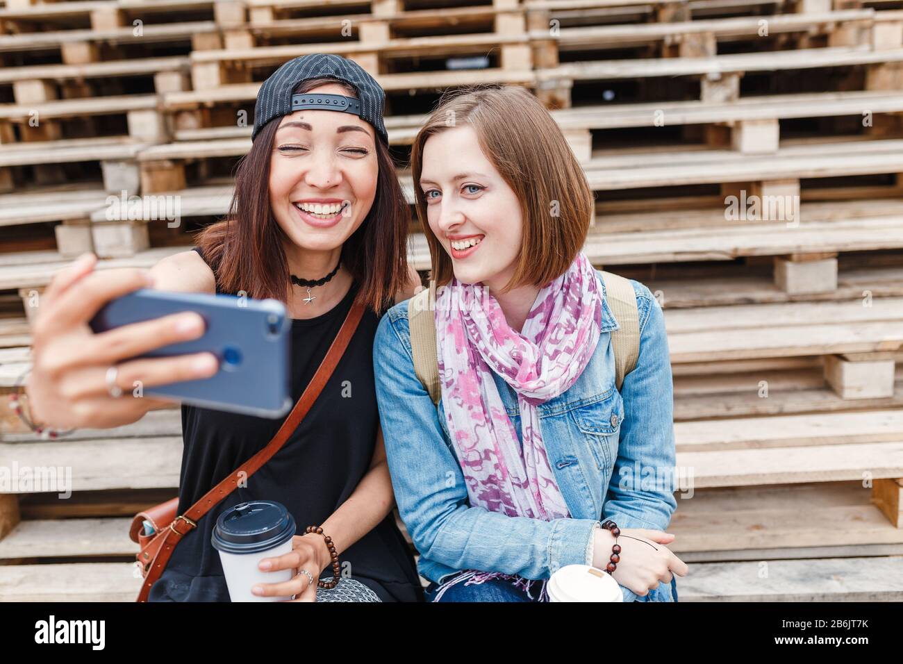 Two young girl students laugh and making selfie on a smartphone while walking outside and drinking coffee, the concept of friendship and fun Stock Photo