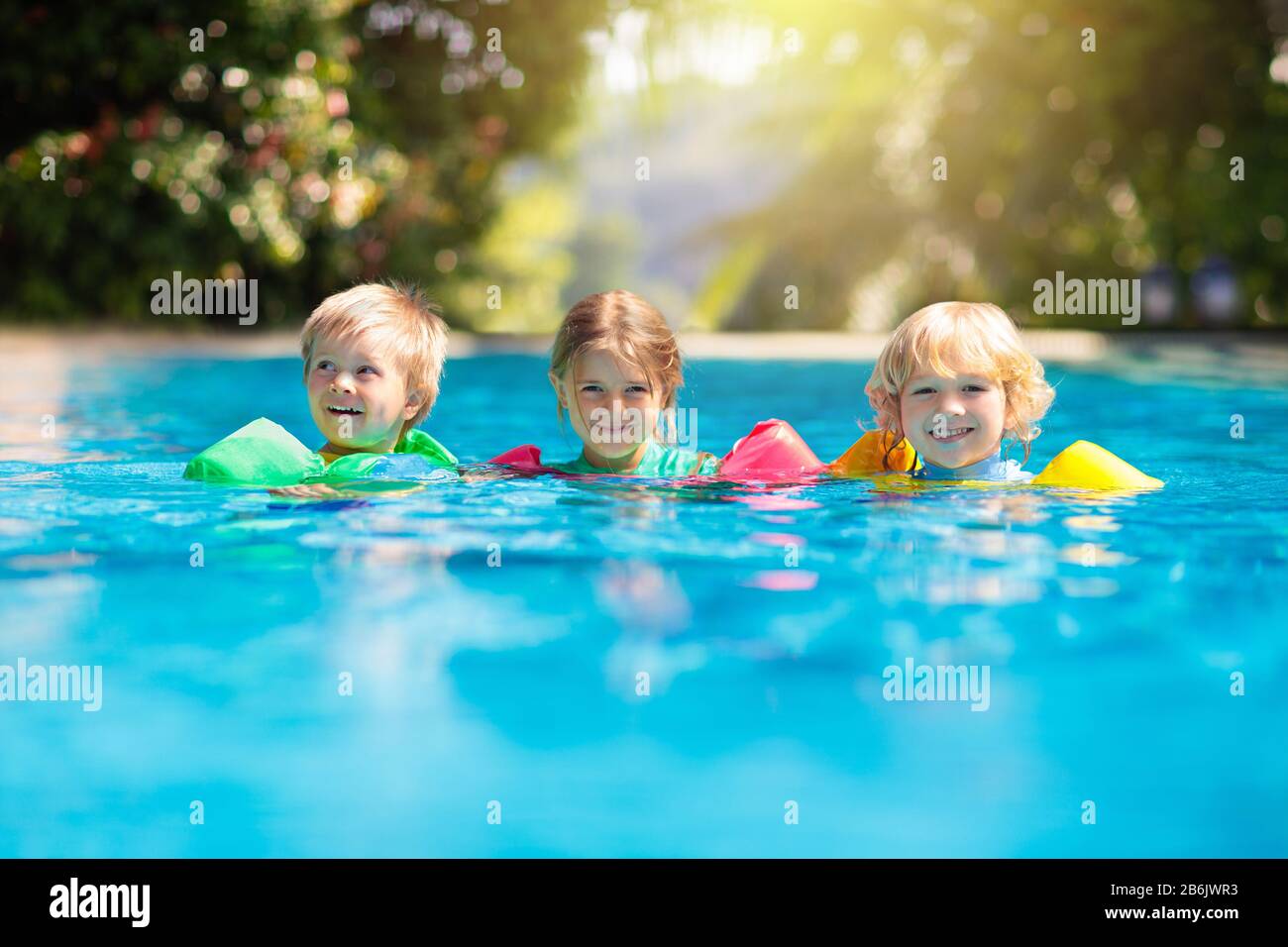Kids play in outdoor swimming pool of tropical resort. Swim aid for young child. Baby learning to dive. Group of children playing in water. Colorful l Stock Photo