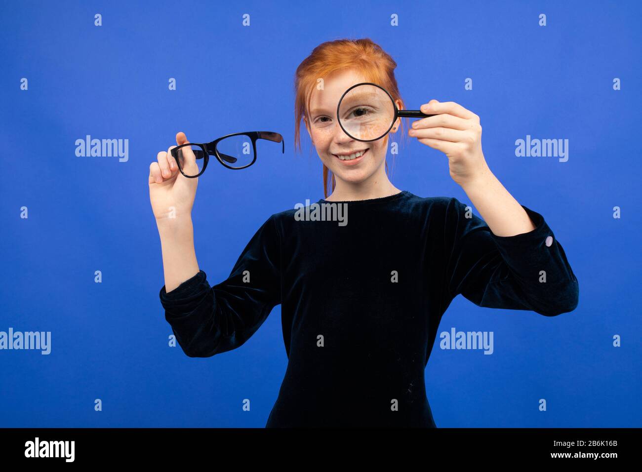 red-haired teenager girl holds glasses for sight and a magnifying glass on a blue background Stock Photo