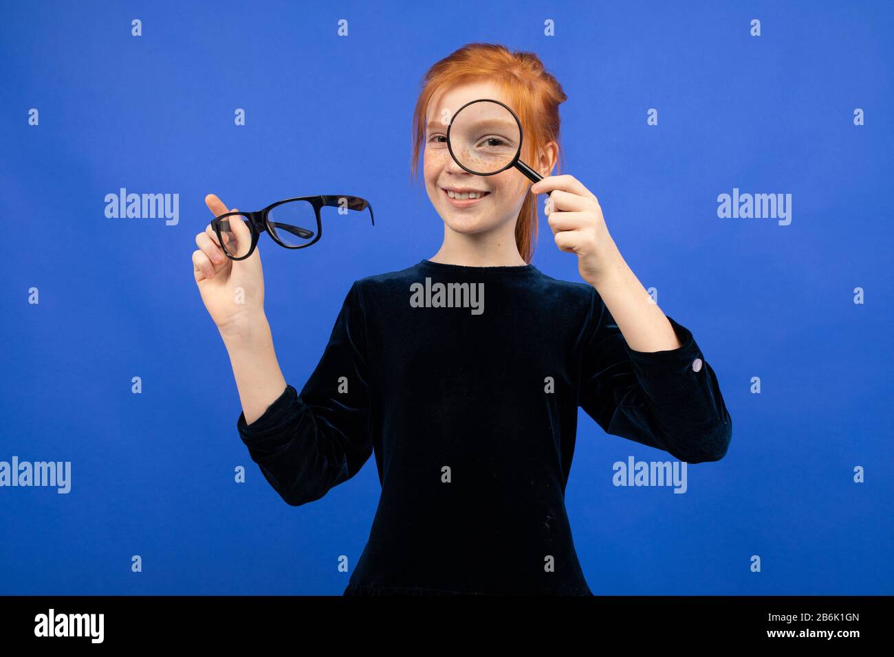 red-haired girl holding glasses for sight and a magnifying glass on a blue background Stock Photo