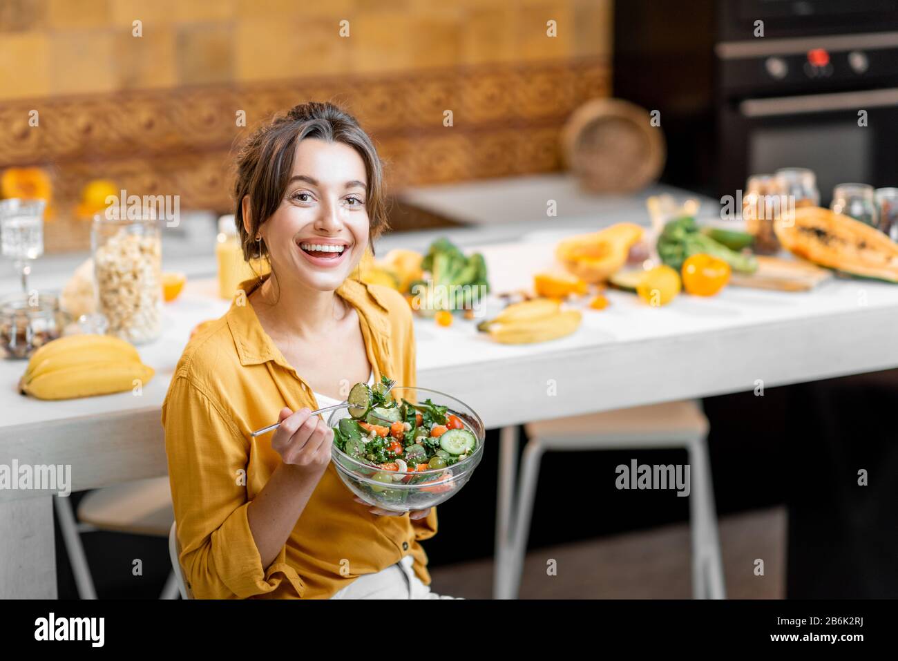 Young and cheerful woman eating salad at the table full of healthy raw vegetables and fruits on the kitchen at home. Concept of vegetarianism, healthy eating and wellness Stock Photo