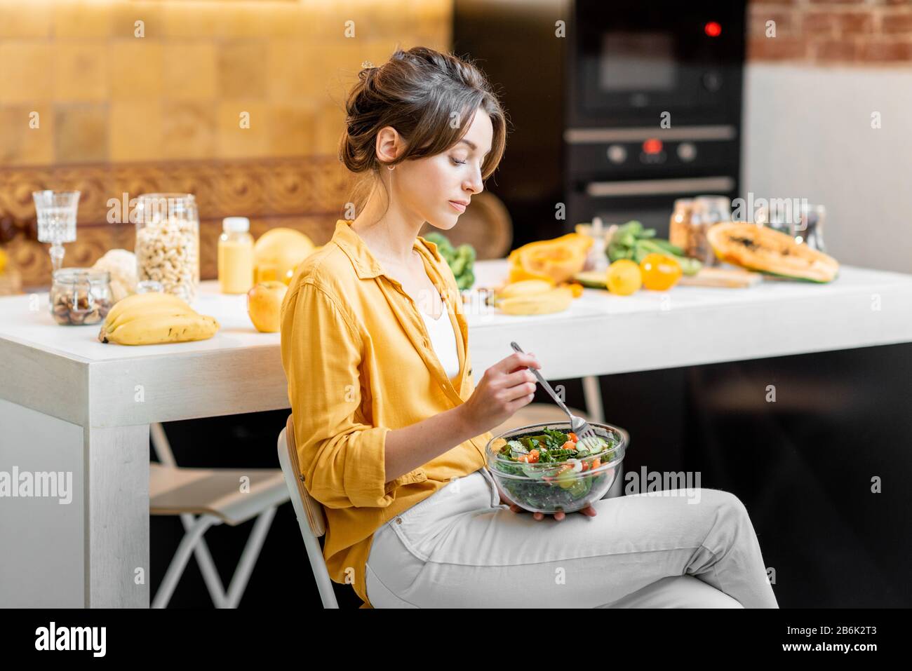 Young and cheerful woman eating salad at the table full of healthy raw vegetables and fruits on the kitchen at home. Concept of vegetarianism, healthy eating and wellness Stock Photo