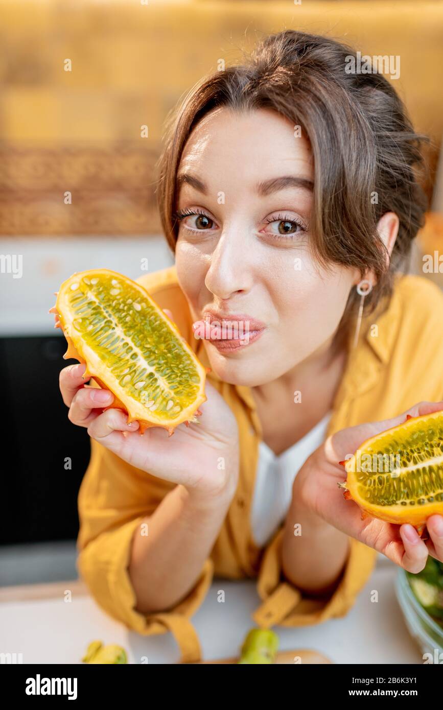 Portrait of a joyful woman with variety of exotic fruits and vegetables on the kitchen, holding sliced cucumis metuliferus. Concept of vegetarianism and healthy eating Stock Photo