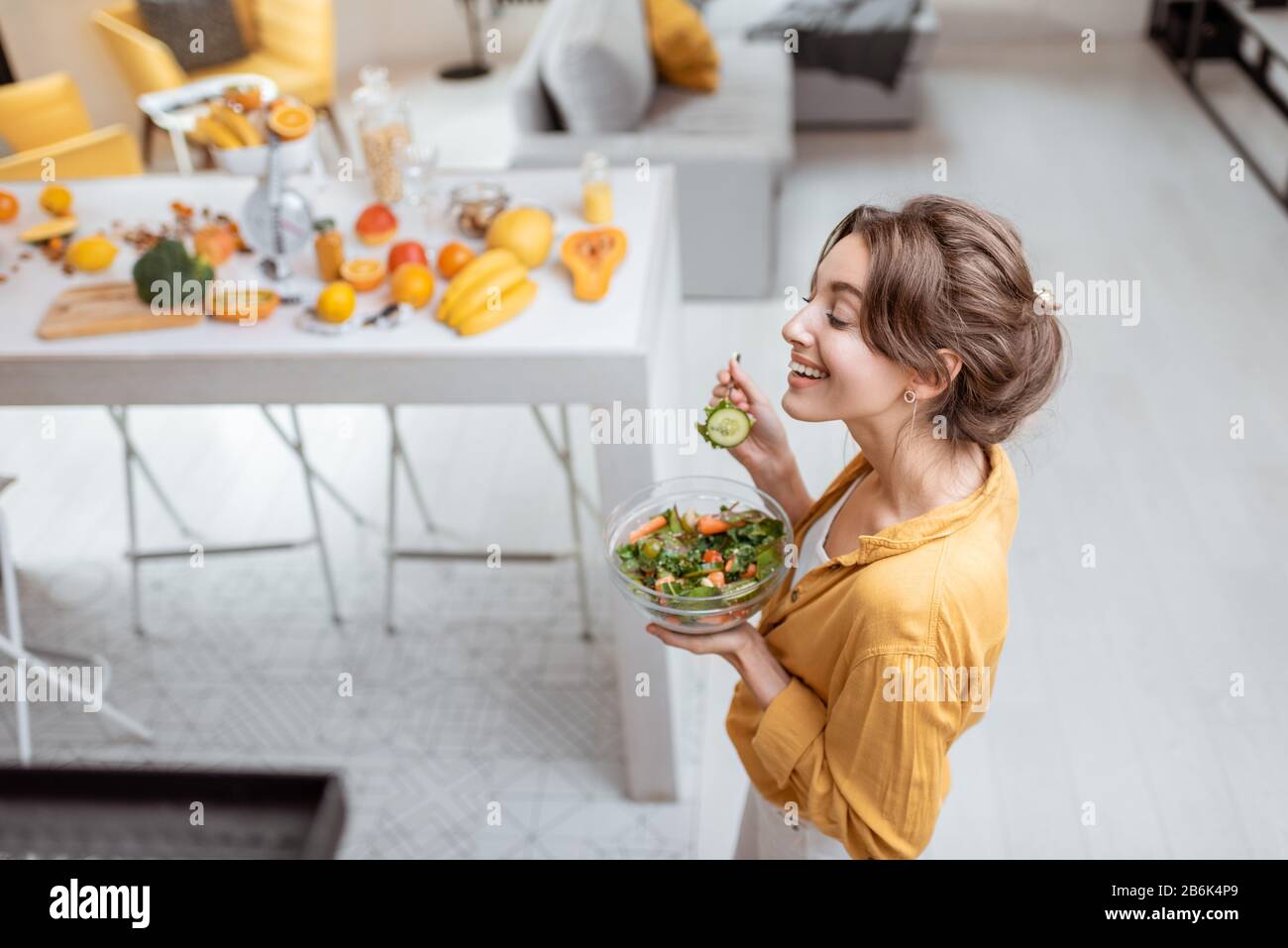Portrait of a young and cheerful woman dressed in bright shirt eating salad at home. Concept of wellbeing, healthy food and homeliness Stock Photo