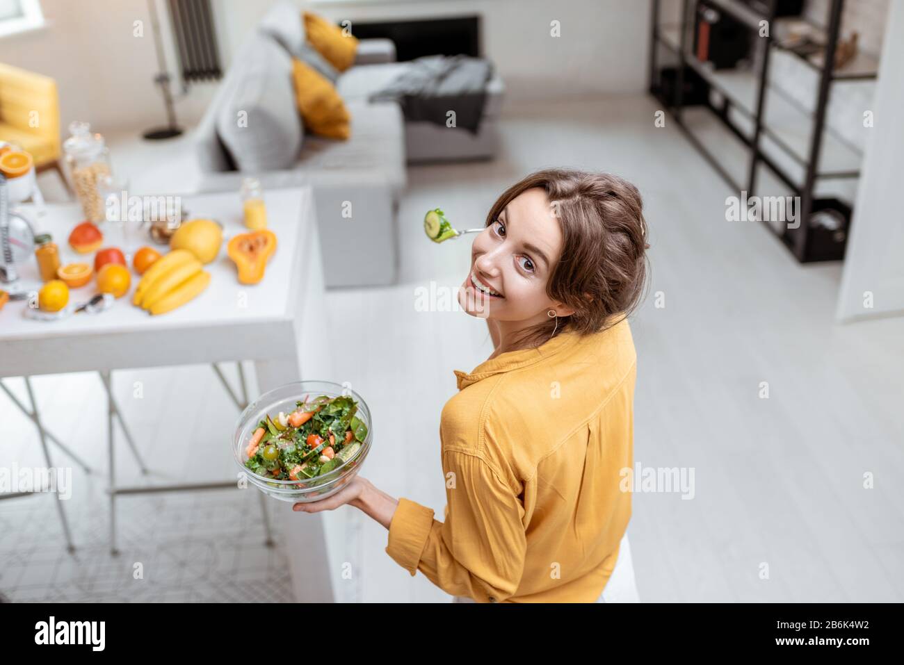 Portrait of a young and cheerful woman dressed in bright shirt eating salad at home. Concept of wellbeing, healthy food and homeliness Stock Photo