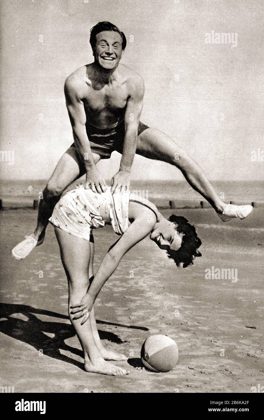Circa 1950's photograph showing a man and a woman  playing leap frog on an English beach, wearing typical  bathing / swimming costumes of the time. Behind can be seen wooden groins which were intended to stop the sands from drifting. Stock Photo