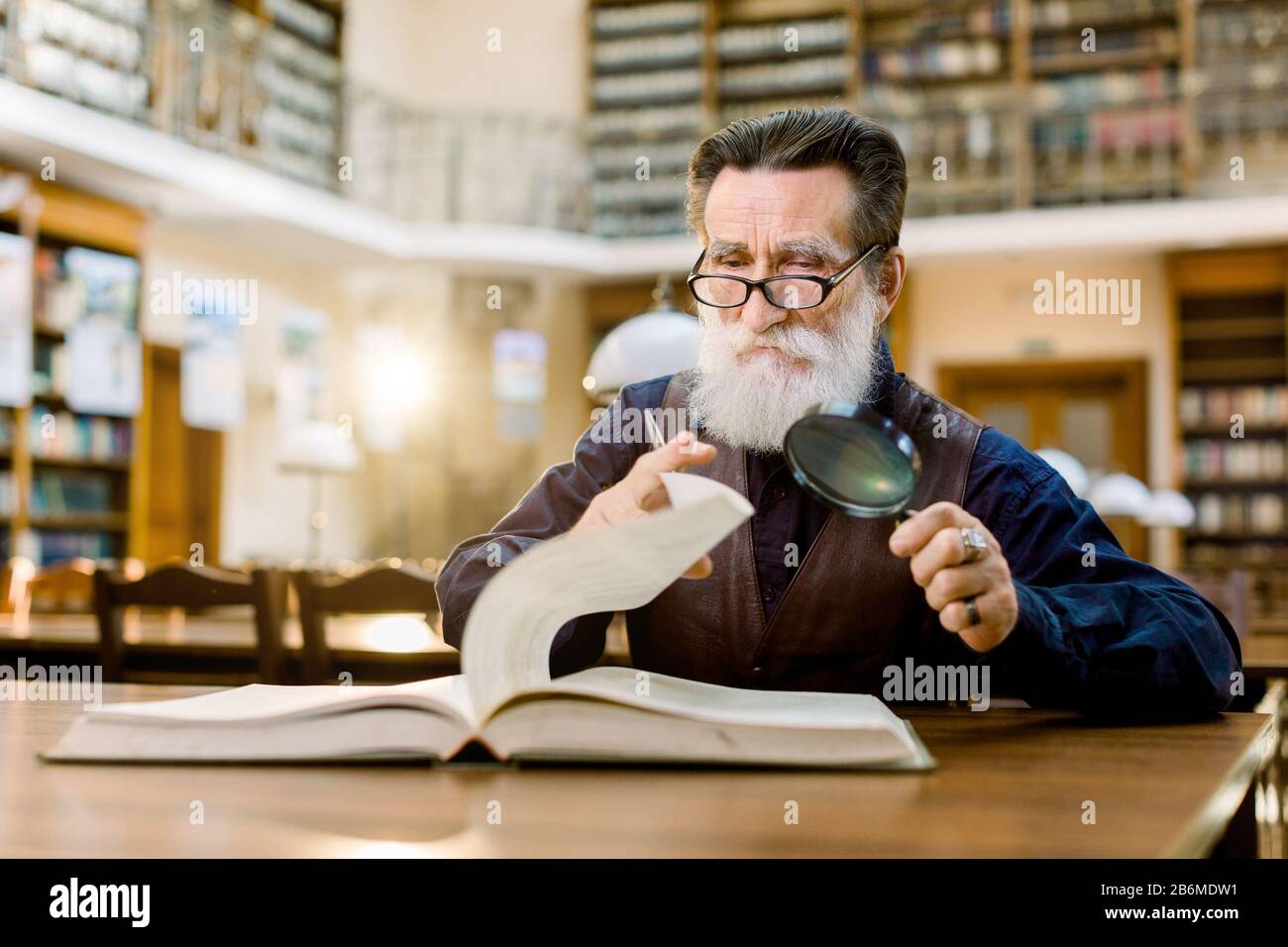 Senior caucasian bearded man in glasses and stylish shirt and leather vest  sitting at the table in vintage library, holding magnifying glass and Stock  Photo - Alamy