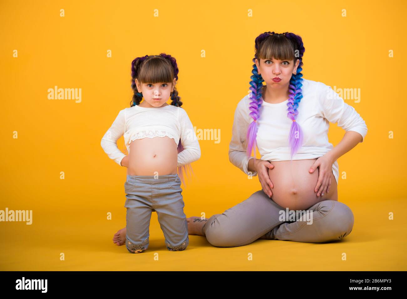 Funny positive mom and daughter show their pregnant belly posing in the studio on a yellow background. The concept of foolishness and family play. Cop Stock Photo