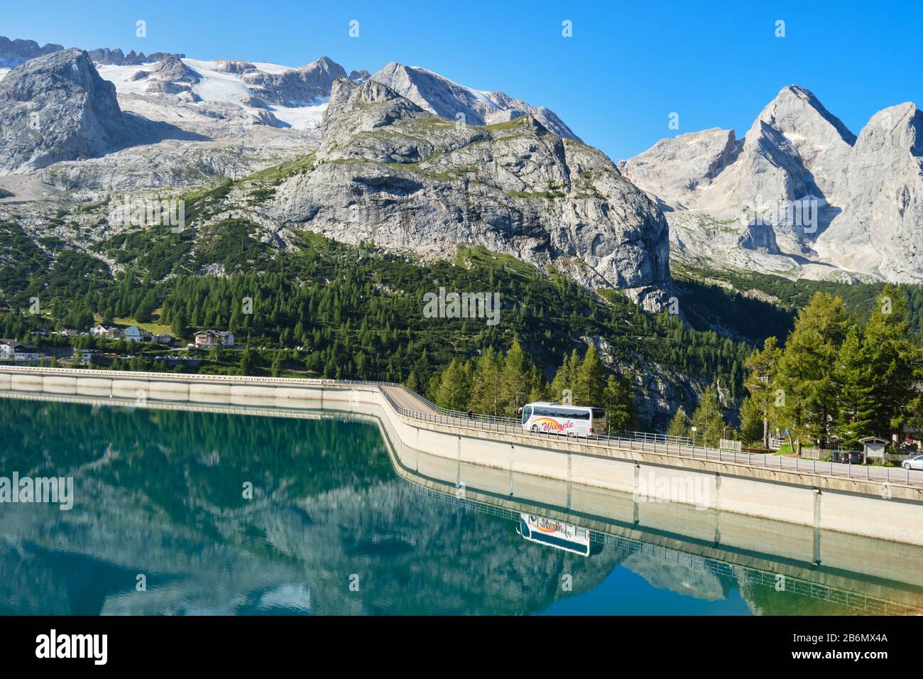 Fedaia Lake, Italy - August 27, 2019: Bus on the Fedaia lake dam, reflected in the turquoise waters, on a bright sunny day, with Marmolada glacier in Stock Photo