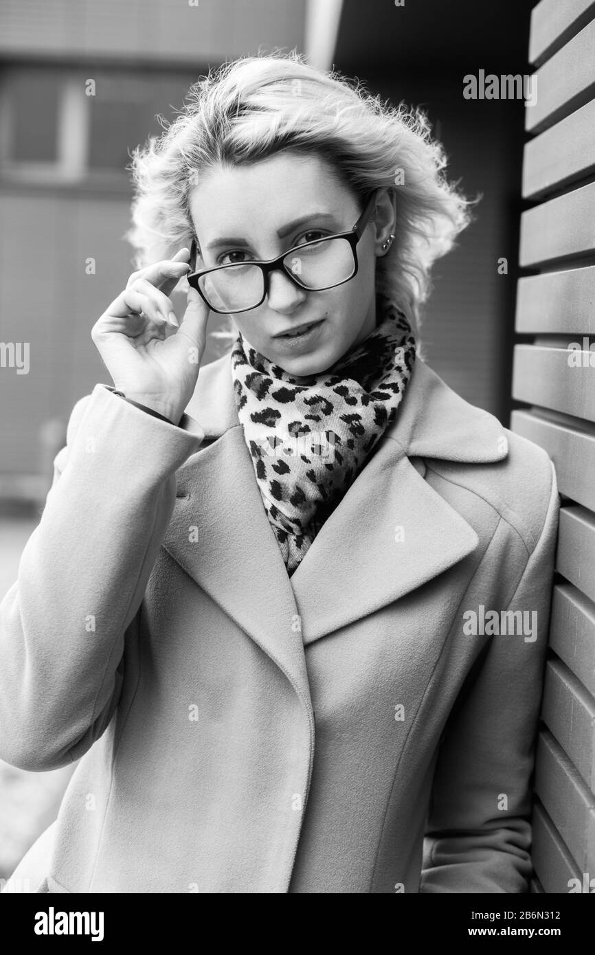 sad blond girl in eyeglasses near wall looking at camera, smiling, monochrome Stock Photo