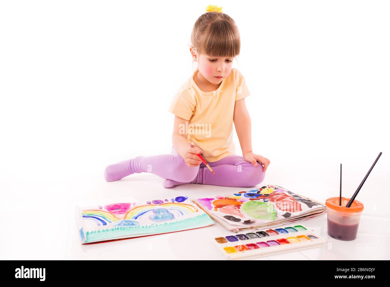 Cute girl with a calm face sitting on the floor with her hand on knee. A child paints with watercolors Stock Photo