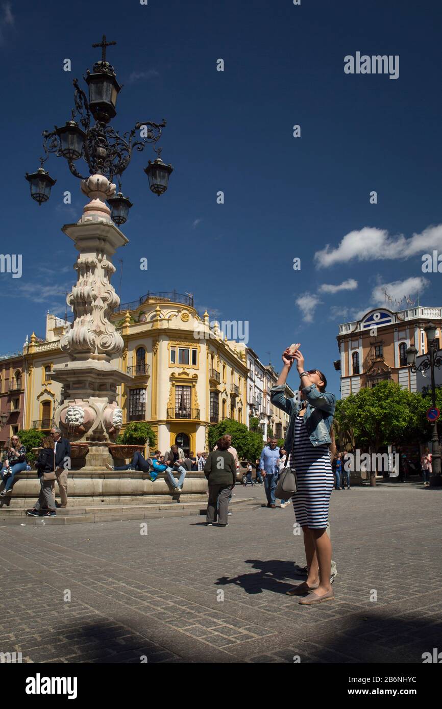 Tourist taking photos with a mobile phone next to the fountain-lamppost located at Virgen de los Reyes Square, Seville, Spain Stock Photo