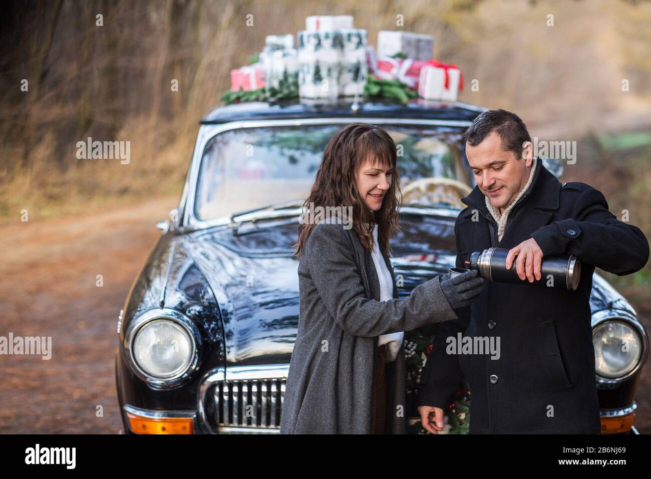 Couple in coats and warm glove holding metal cup pouring hot tea from thermos during cold day by the retro car with Christmas presents Stock Photo