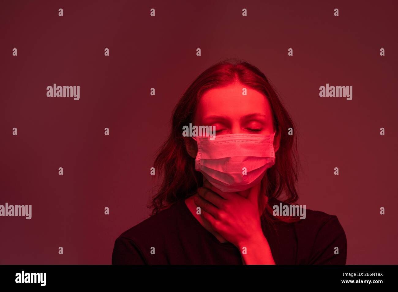 Infected young woman in face mask keeping eyes closed and feeling pain in throat, dark red light