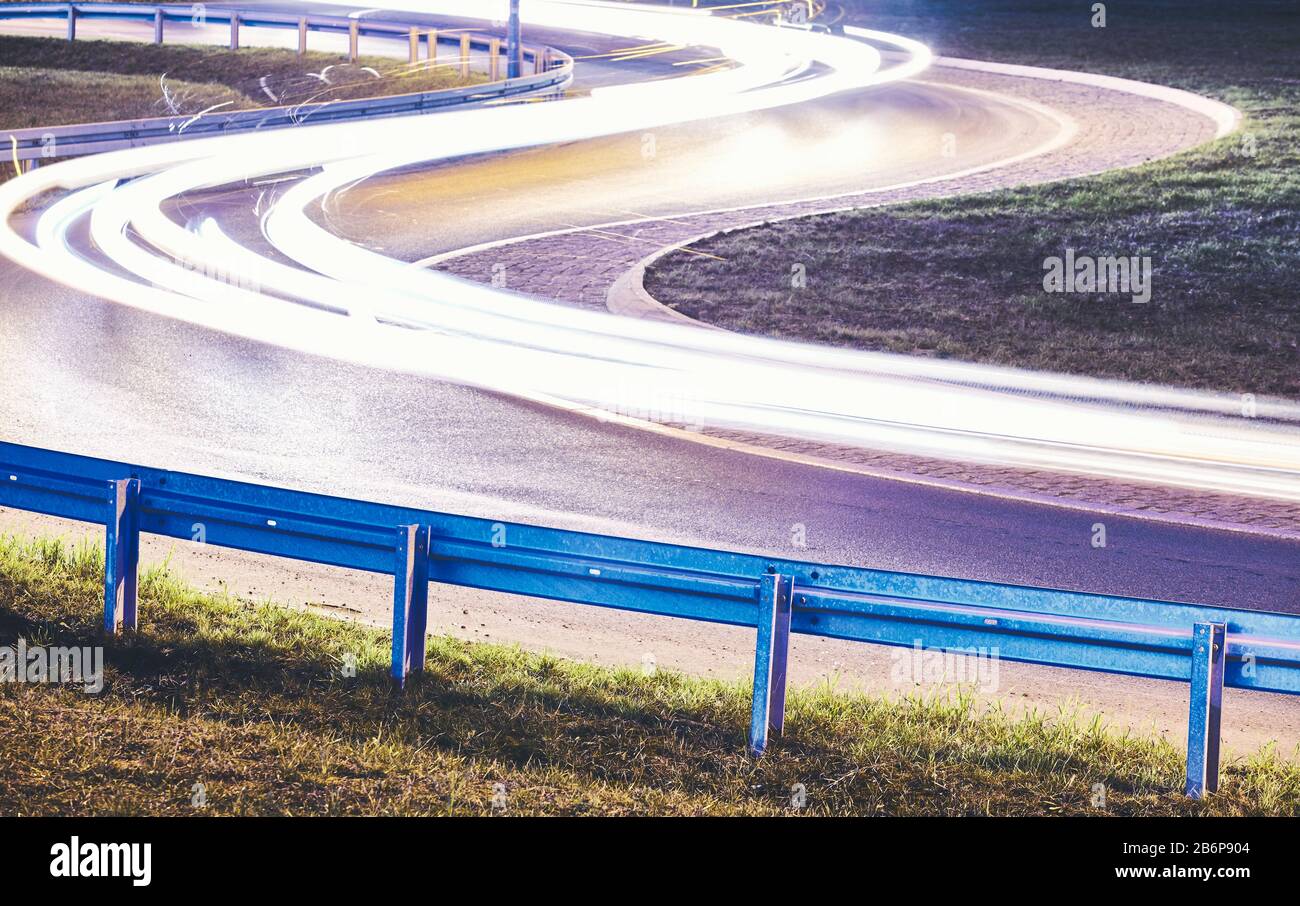 Curvy road with car light trails at night, focus on guardrail, color