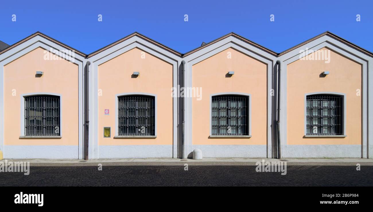 Old-fashioned factory shed with a pale pink facade on a blue sky, zig-zag roof, hydrant and windows in the southern suburbs of Milan, Italy Stock Photo