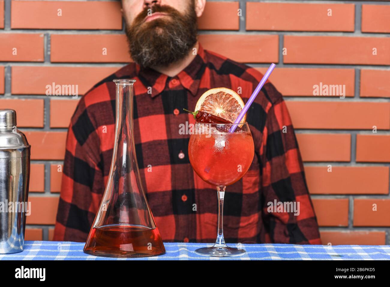 Man in checkered shirt on brick wall background prepares drinks. Alcoholic drinks concept. Barman with long beard and mustache on serious face made alcoholic cocktail with orange Stock Photo
