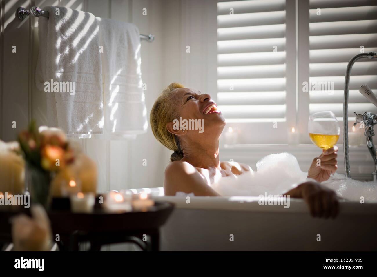 Mid-adult woman relaxing in a bubble bath with a glass of wine and surrounded by candles. Stock Photo