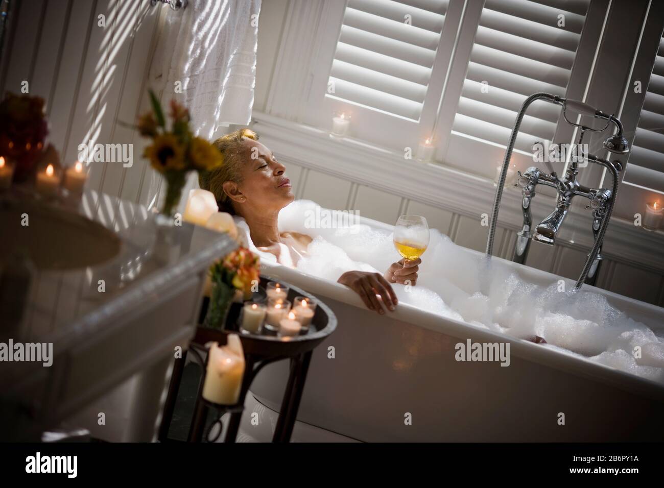 Mid-adult woman relaxing in a bubble bath with a glass of wine and surrounded by candles. Stock Photo