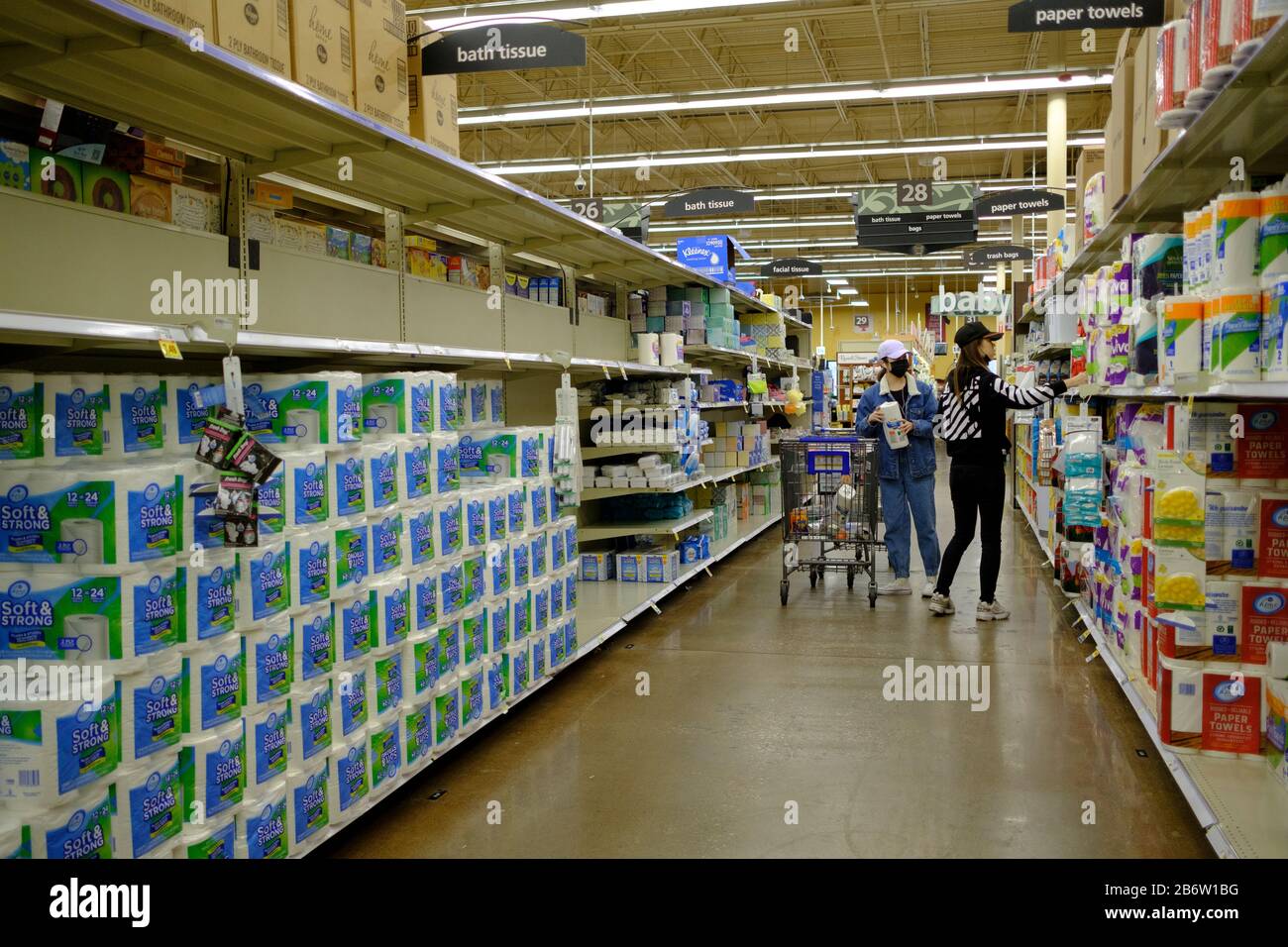 03112020 - Bloomington, Indiana, USA: A woman wearing a mask shops for paper towels near the toilet paper section at a Kroger in Bloomington, Ind., where shoppers were stocking up on toilet paper, and other items on the day World Health Organization declared Coronavirus to be a pandemic. Toilet paper, wipes, protective breathing masks, and other items are either sold out at local stores, or are in short supply. Stock Photo